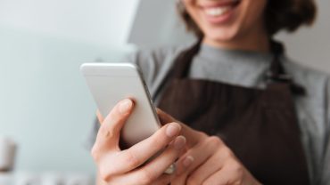 Close up portrait of a younf smiling woman in apron using mobile phone indoors